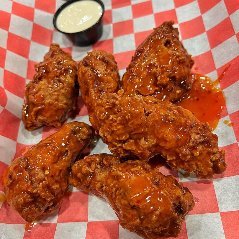 Six hot chicken wings with sauce and a side of ranch dressing on red and white checkered paper.