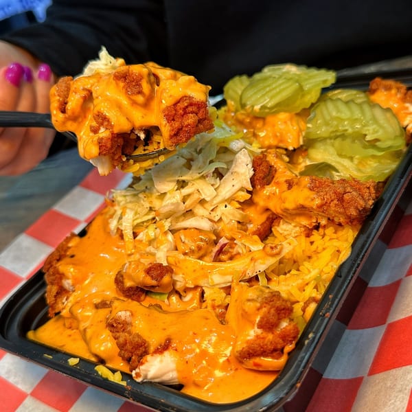 A fork lifts a piece of hot chicken from a tray of rice, coleslaw, and pickles.